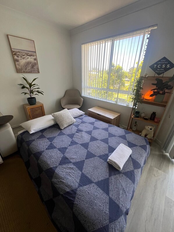 Un dormitorio con cama, una silla, una mesita de noche, una ventana con persianas, una planta en maceta, un oso de peluche y un cuadro en la pared.