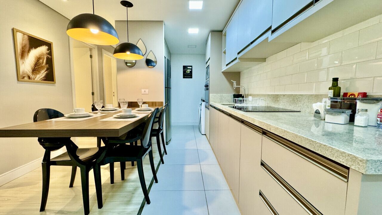 A modern kitchen with a dining area, featuring a long table, chairs, pendant lights, and a tiled backsplash.