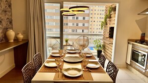 Un coin repas avec une grande table dressée pour six personnes, une cuisine dotée d’un four moderne et une vue sur un immeuble de grande hauteur, visible à travers de larges fenêtres.