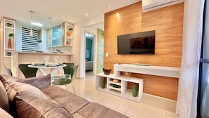 Ein modernes Wohnzimmer mit einer Couch, einem Esstisch mit Stühlen, einem an einer Holzwand montierten Fernseher und einer Küchenzeile im Hintergrund.