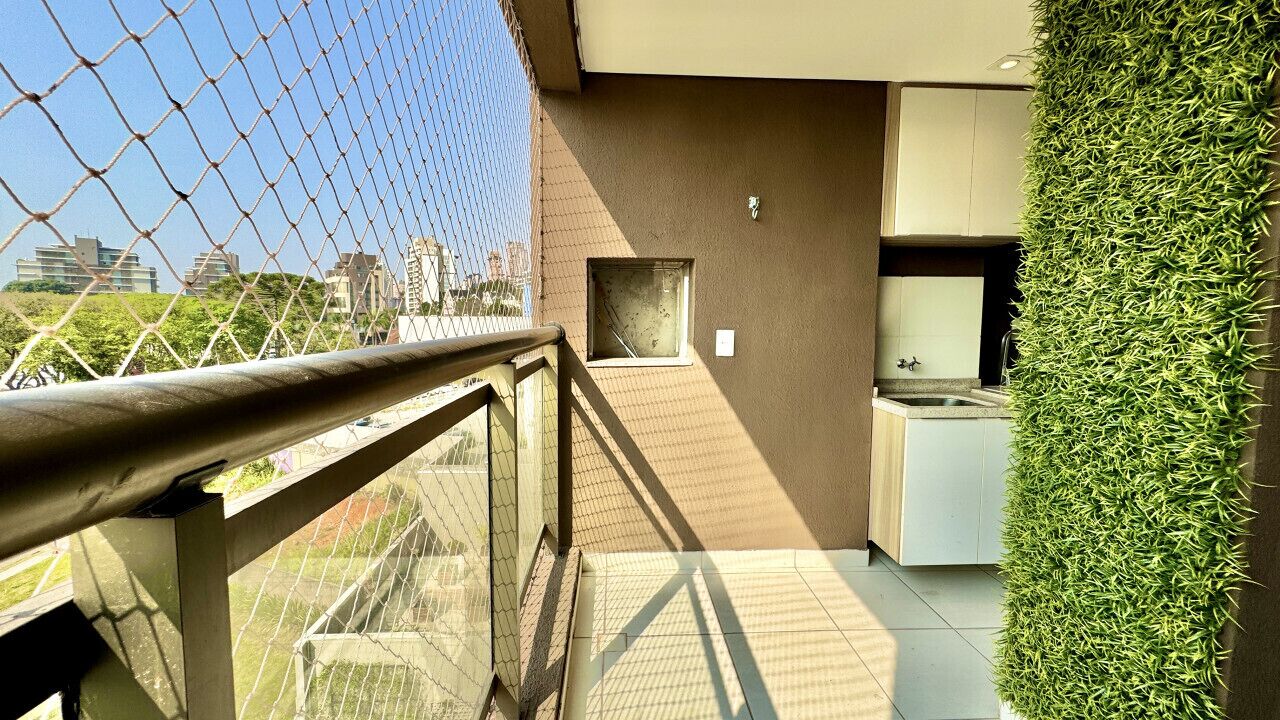 A balcony with a chain-link fence, a small window, and a kitchen area with cabinets and a sink.