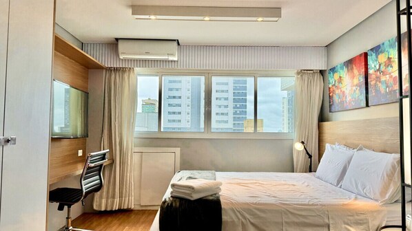 A modern hotel room with a large bed, a desk with a chair, a window with curtains, and a city view. - Premium Studio Lifespace Curitiba Batel com vaga (Curitiba)