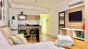 A modern hotel room with a bed, a small kitchenette, a dining area, and a TV mounted on the wall.