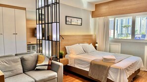 A modern hotel room with a bed, a sofa, a TV, and a window with a city view.