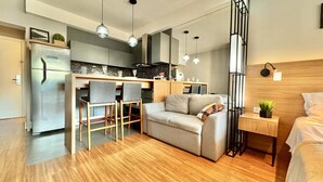 A modern kitchen with a built-in refrigerator, wooden dining table, and bar stools. There is a sofa and a small bedside table with a plant. - Luxo e Estilo juntos nesse Studio no Lifespace CWB (Curitiba)