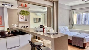 Une chambre d’hôtel moderne dotée d’une kitchenette, d’un coin repas, d’un lit et d’une grande fenêtre offrant une vue sur la ville.
