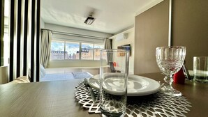 Une chambre d’hôtel avec une table à manger dressée pour deux personnes, un vase en verre transparent et une vue sur la ville par la fenêtre.