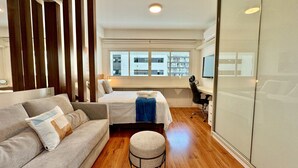 A modern hotel room with a sofa, a bed, a desk, and a large window offering a city view. - Studio Luxo na frente do Shopping Curitiba, c/vaga (Curitiba)