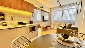 Uma cozinha moderna com micro-ondas embutido, pia e bancada. Há uma área de refeições com uma mesa posta para dois, um sofá e uma TV ao fundo.
