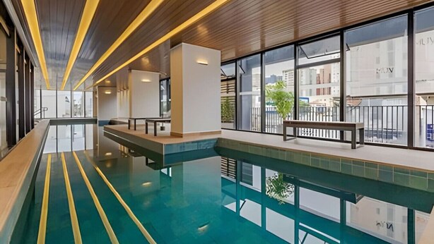 A modern indoor swimming pool with clear glass walls, wooden ceiling, and a bench by the pool.