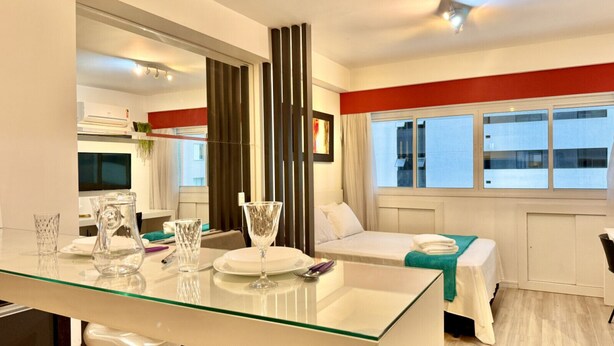 A hotel room with a glass dining table, a bed with a teal bedspread, a window with a view of buildings, and a large mirror.