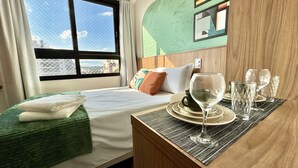 A hotel room with a bed, a small table with glasses and a cup, and a window with a view of buildings. - Studio compacto próximo CoutoPereira com Ar e vaga (Curitiba)