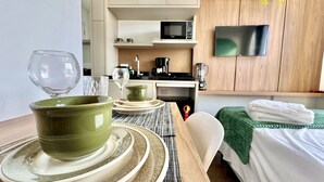 A hotel room with a dining table set with plates and bowls, a bed with a green blanket, a TV mounted on the wall, and a microwave in the kitchenette. - Studio compacto próximo CoutoPereira com Ar e vaga (Curitiba)