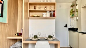 A compact kitchen with wooden shelves, a dining table set with green cups and wine glasses, and a small refrigerator. - Studio compacto próximo CoutoPereira com Ar e vaga (Curitiba)