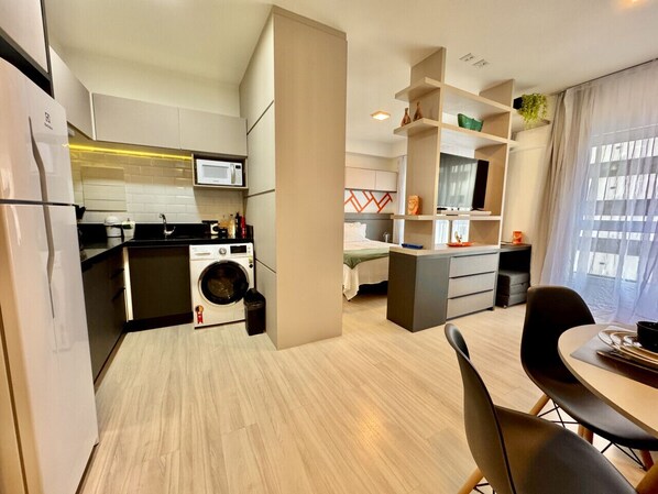 A modern kitchen with a washing machine, a dining area with a table and chairs, and a bed in the background.