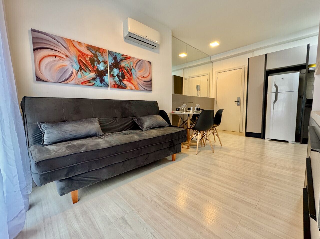 A modern living room with a dark grey sofa, a dining table with chairs, and a kitchen area with a refrigerator.