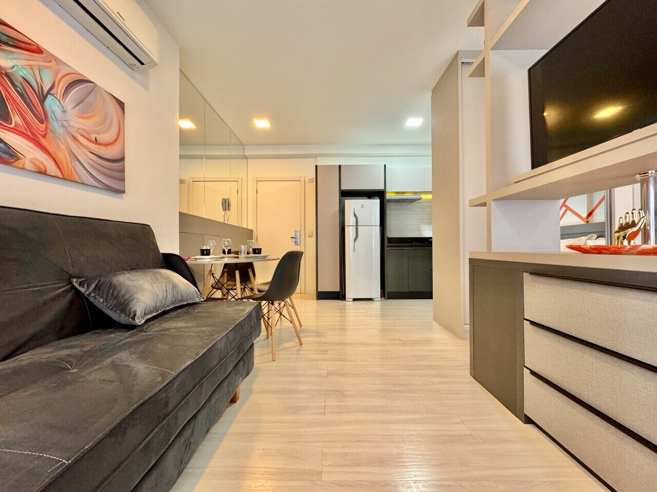 A modern living room with a sofa, a dining area with chairs, a kitchen with a refrigerator, and a TV mounted on the wall.