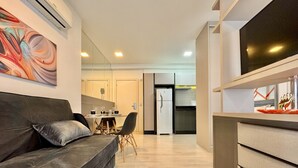 Una sala de estar moderna con un sofá, un comedor con sillas, una cocina con refrigerador y un televisor montado en la pared.