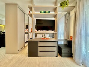 A modern hotel room with a central island, a bed with white linens, a TV on the wall, and a staircase leading to an upper level.