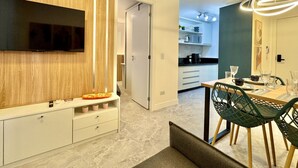 A modern kitchen with a built-in TV, a dining area with green chairs, and a hallway leading to a bedroom.