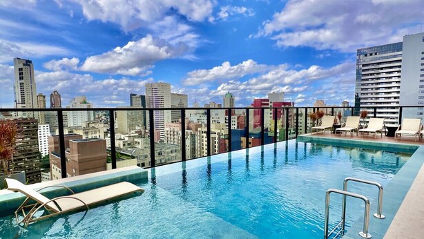 A rooftop infinity pool with lounge chairs, a city skyline view, and a clear blue sky.