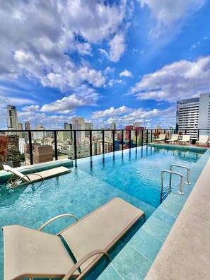 Una piscina infinita en la azotea con sillones de descanso, vistas del horizonte urbano y un cielo azul despejado.