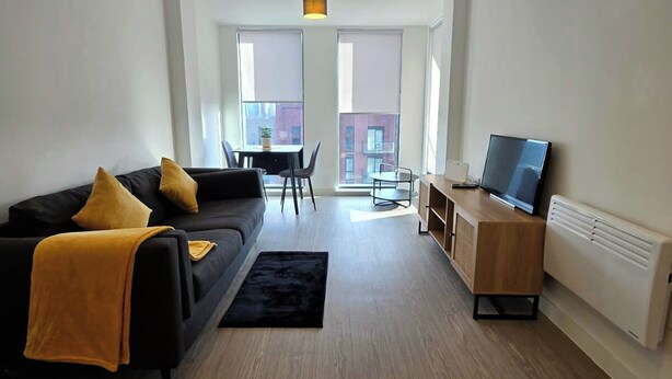 Ein modernes Wohnzimmer mit einem schwarzen Sofa, gelben Kissen, einem schwarzen Teppich, einem Esstisch mit Stühlen und einem Fernseher auf einer Holzkassette.