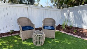 Un jardin avec deux fauteuils en osier dotés de coussins bleus, une petite table ronde et une clôture blanche.