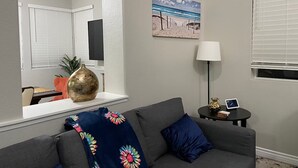 Un salon avec un canapé gris, un coussin bleu, un vase décoratif, un tableau représentant une plage et une petite table sur laquelle se trouvent une lampe et une horloge.