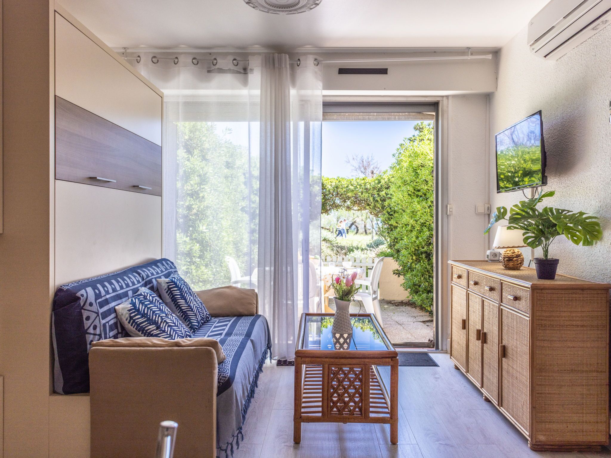 A modern living room with a sofa, a coffee table, and a cabinet. There is a sliding glass door leading to an outdoor area with greenery.