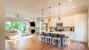 A modern kitchen with white cabinets, a central island with stools, a built-in oven, and a flat-screen TV.