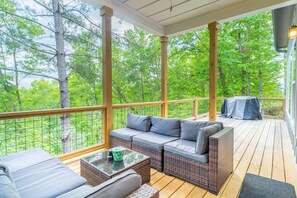 Eine überdachte Terrasse im Freien mit einem Rattan-Sofa und einem Glastisch, umgeben von Holzsäulen und großen Fenstern mit Blick auf einen Wald.