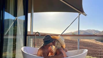 A couple in a large white bathtub on a wooden deck with a view of a hilly landscape.