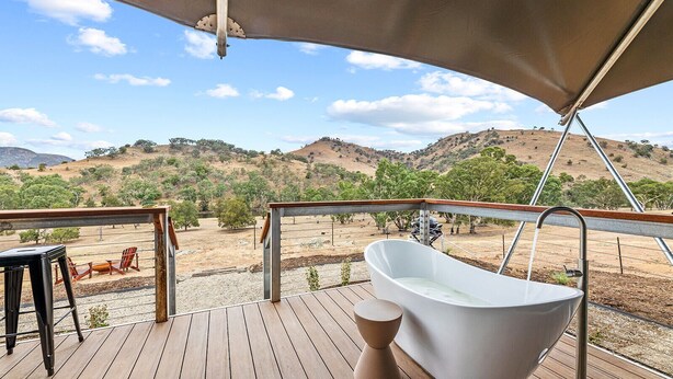 Une terrasse en bois avec une baignoire blanche autoportante, une chaise en bois et une rambarde en métal donnant sur un paysage vallonné.