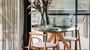 Un comedor con mesa de vidrio, sillas de madera y un florero con plantas secas.