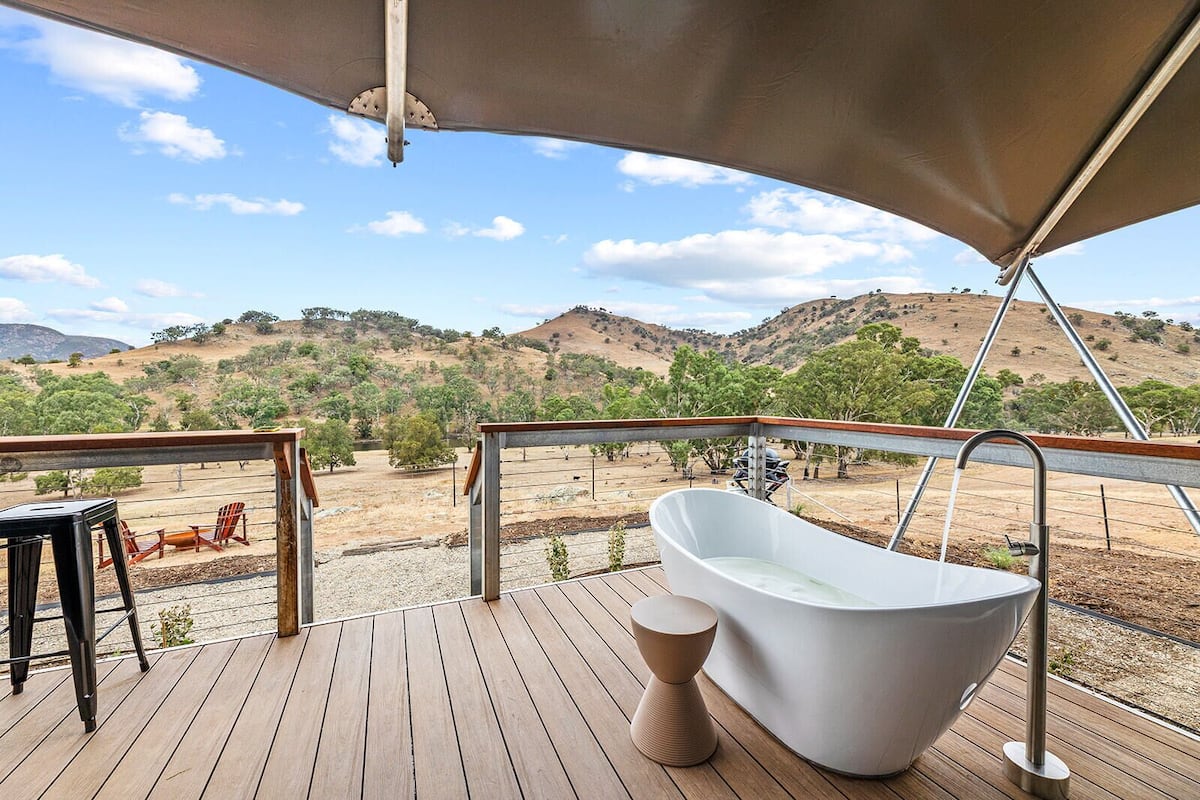 A wooden deck with a white freestanding bathtub, a wooden chair, and a metal railing overlooking a hilly landscape.