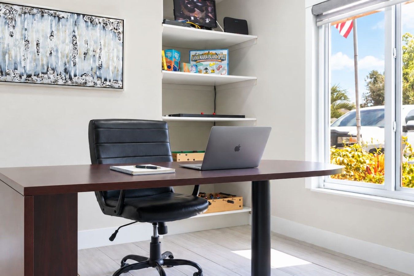 A modern office space with a desk, chair, laptop, shelves with board games, and a large window with a view of parked cars and greenery.