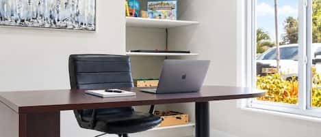 A modern office space with a desk, chair, laptop, shelves with board games, and a large window with a view of parked cars and greenery.