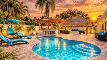 Ein Swimmingpool mit Liegestühlen und einer Grillanlage, umgeben von Palmen und einem Sonnenuntergang.