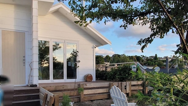 A white house with a front porch, wooden steps, and a garden area with plants and a white Adirondack chair.