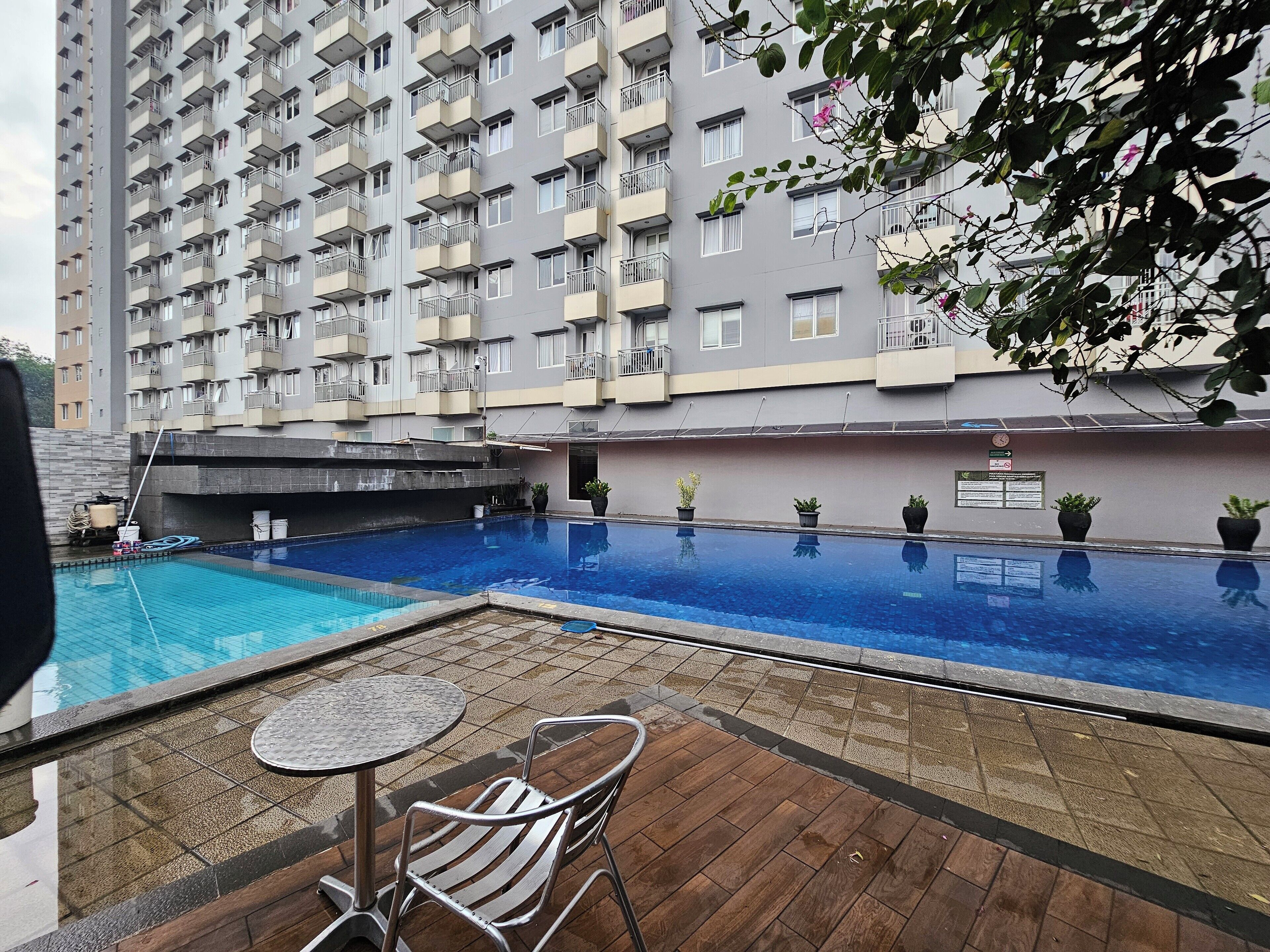 A swimming pool with a wooden deck and metal chairs, surrounded by a high-rise building with balconies.