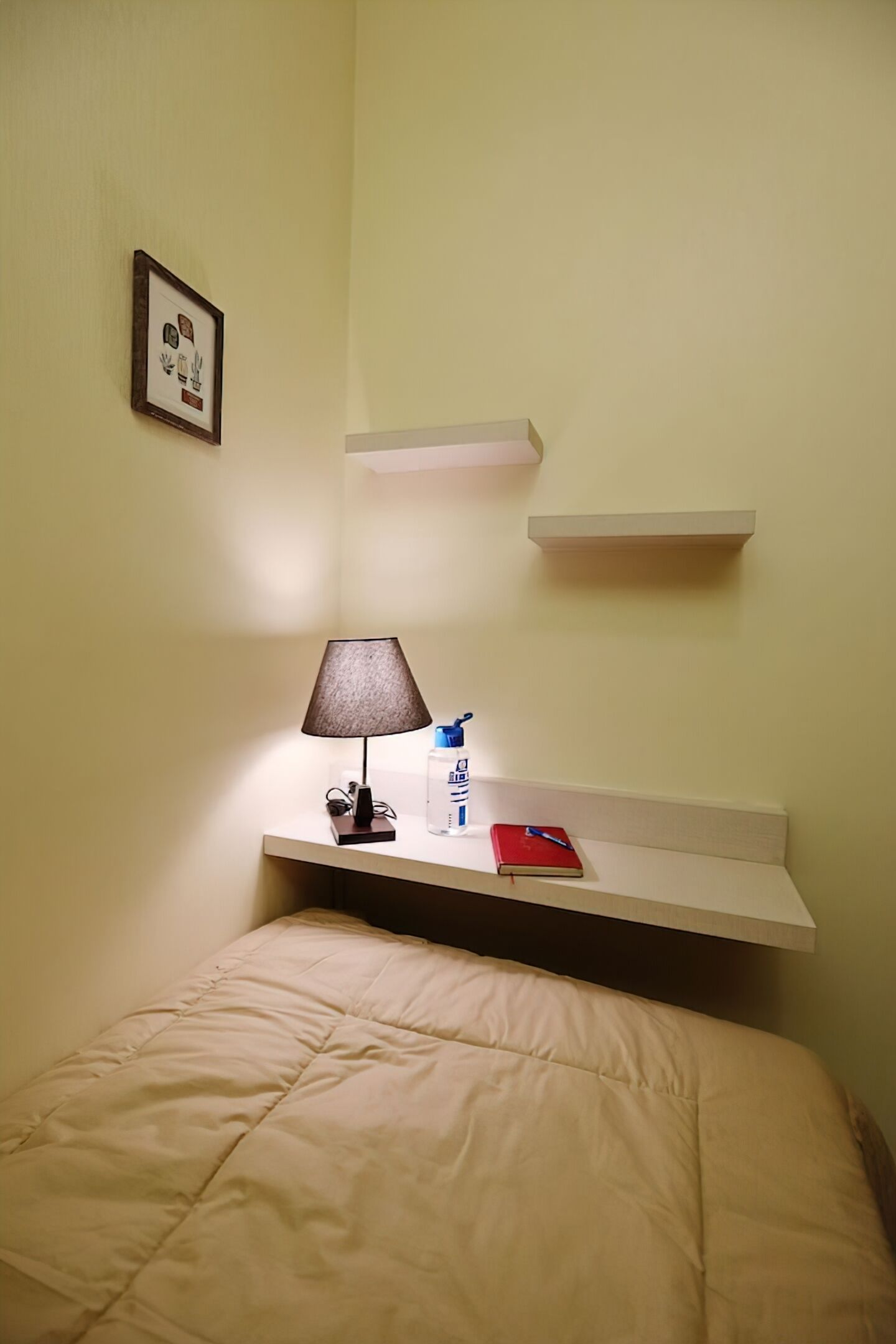 A hotel room with a bed, a bedside lamp, a small table with a red notebook, a water bottle, and a framed picture on the wall.
