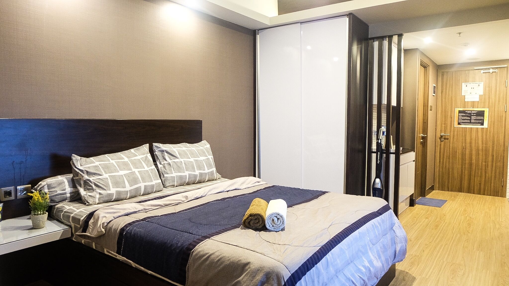 A hotel room with a bed, a nightstand, a potted plant, and a door leading to a bathroom.