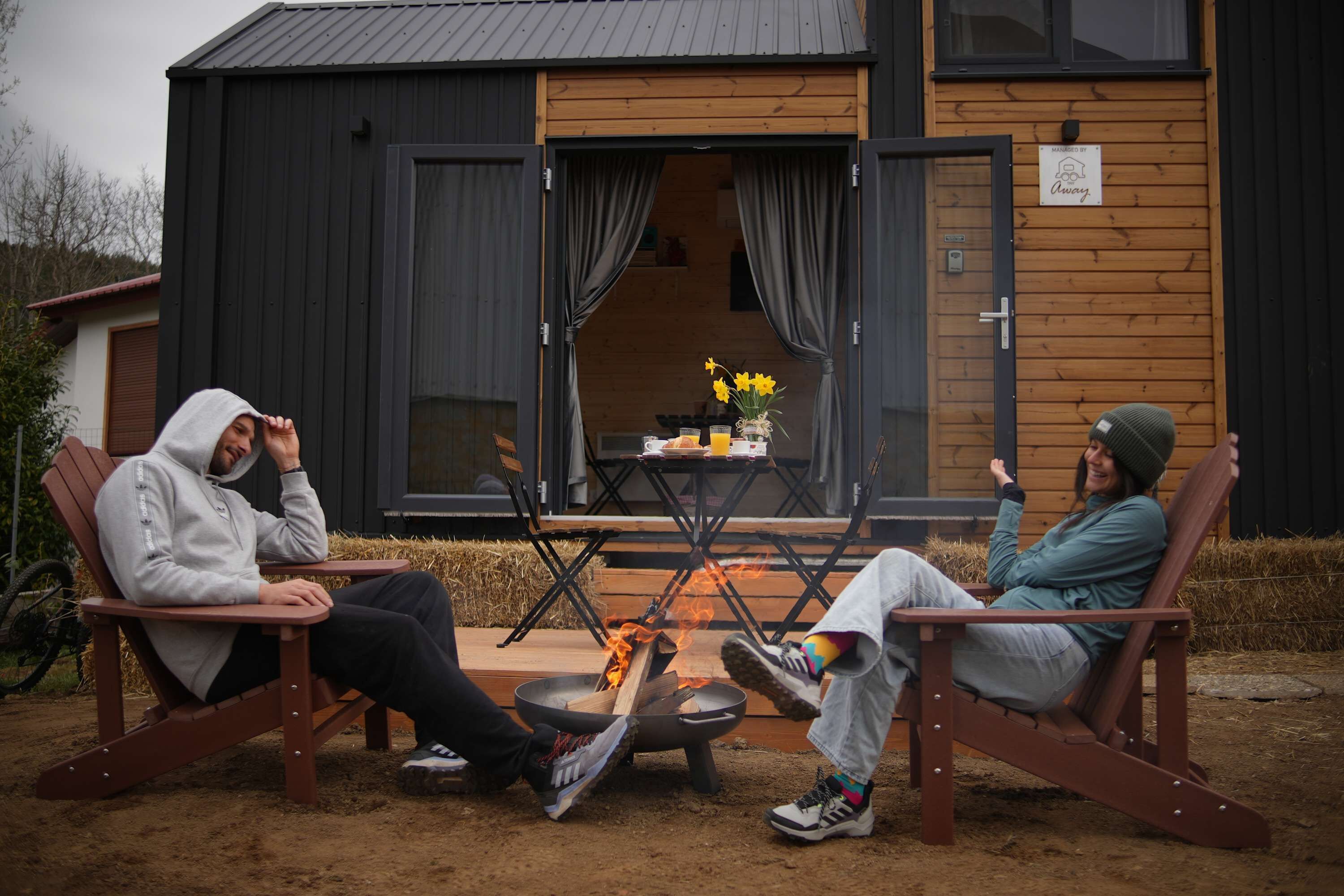 Deux personnes sont assises sur des chaises en bois, à l’extérieur d’un bâtiment, et un foyer est installé entre elles.