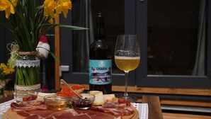 Una tabla de embutidos con carnes, quesos y salsas, una botella de vino Chiaria, una copa de vino y flores amarillas en un jarrón.