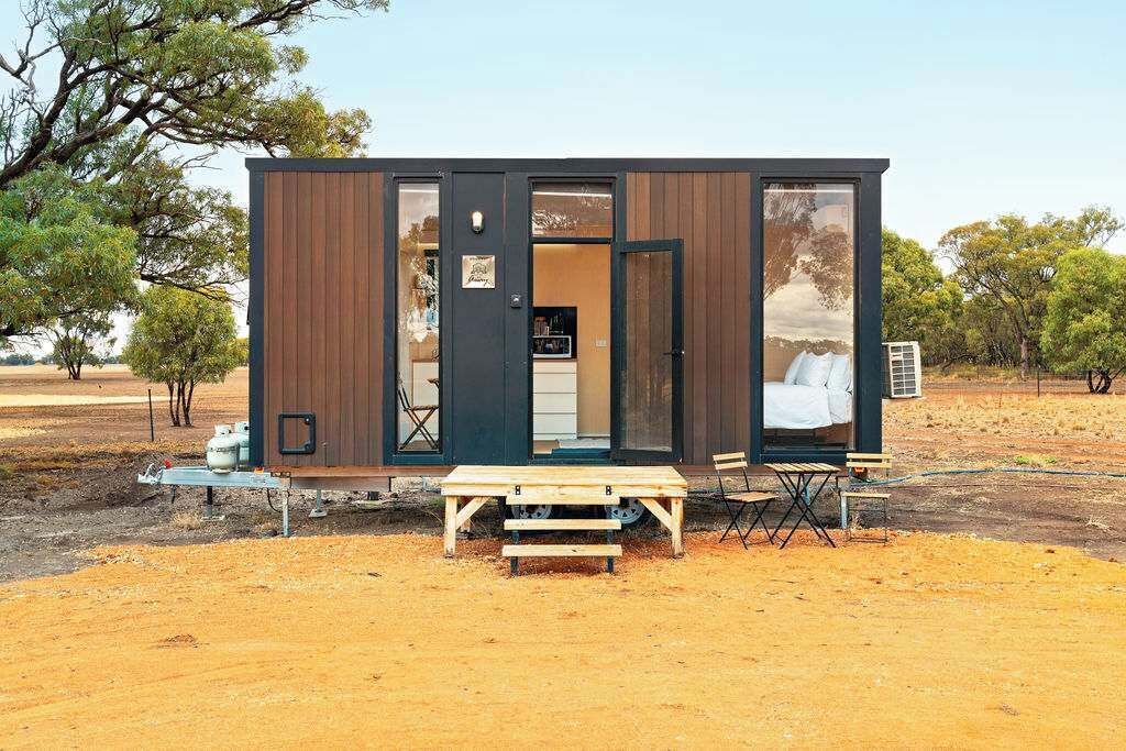 A small, modern cabin with a wooden exterior and large windows, situated in a dry, open landscape with sparse trees.