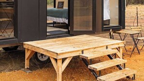 A modern, compact mobile home with a large sliding door, a small outdoor seating area, and a visible bed inside.