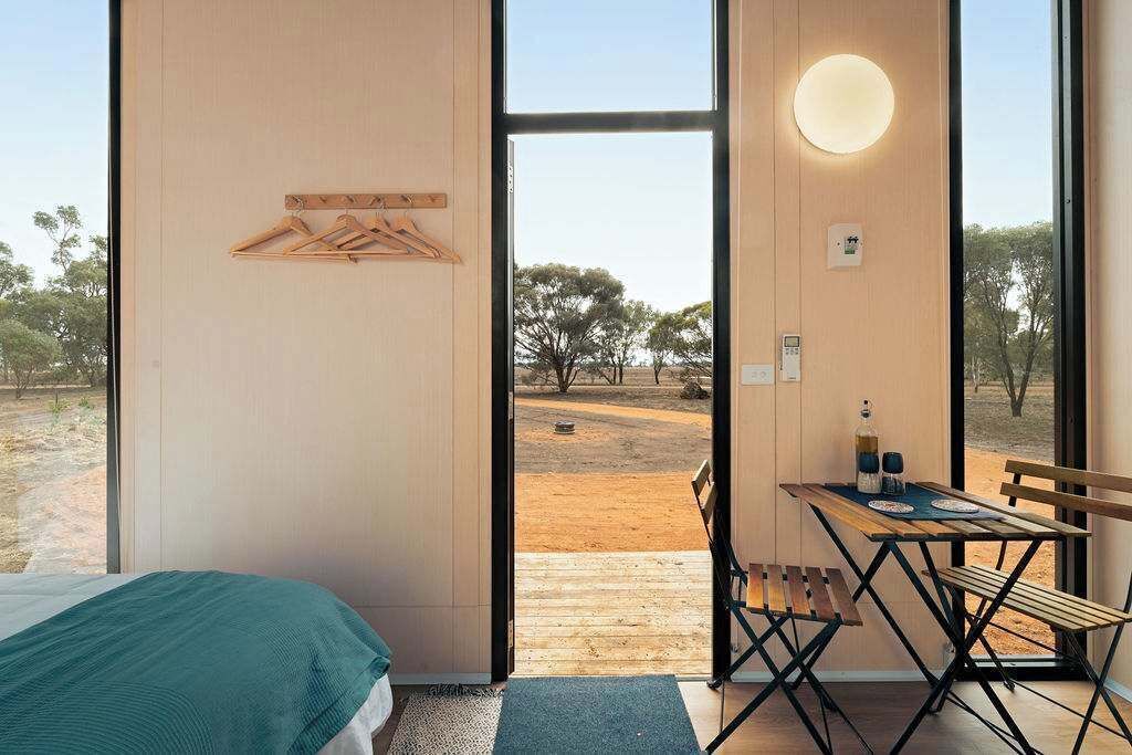 A room with a bed, a table with chairs, a hanging clothes rack, and a large window with a view of trees and a dirt ground.