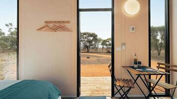 A room with a bed, a table with chairs, a hanging clothes rack, and a large window with a view of trees and a dirt ground.