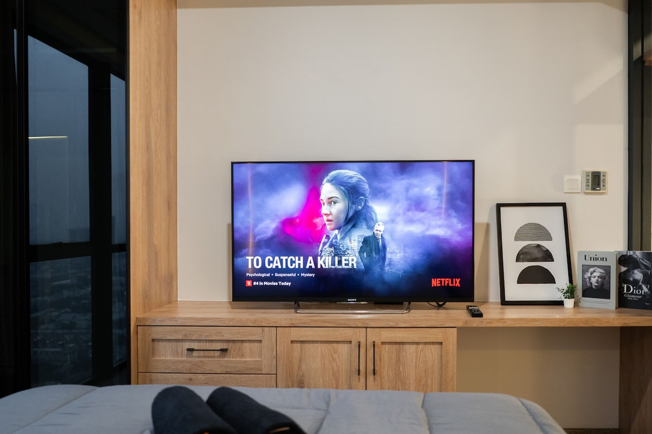 A television displaying a movie titled 'To Catch a Killer' on Netflix, placed on a wooden cabinet in a room with a bed in the foreground.